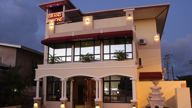 Een hotel met een opvallend bord waarop "Grand Diamond Hotel" staat, bestaande uit een gebouw van twee verdiepingen met een overdekte ingang en een decoratieve fontein in de binnenplaats.