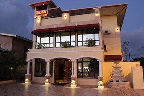 Un hôtel reconnaissable à son enseigne imposante indiquant « Grand Diamond Hotel », avec une façade à deux étages, une entrée couverte et une fontaine décorative dans la cour.