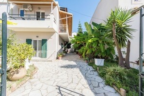 A narrow stone-paved courtyard with potted plants and a small tree, leading to a white building with balconies and a door. - Cavos Beach House (Corfu)
