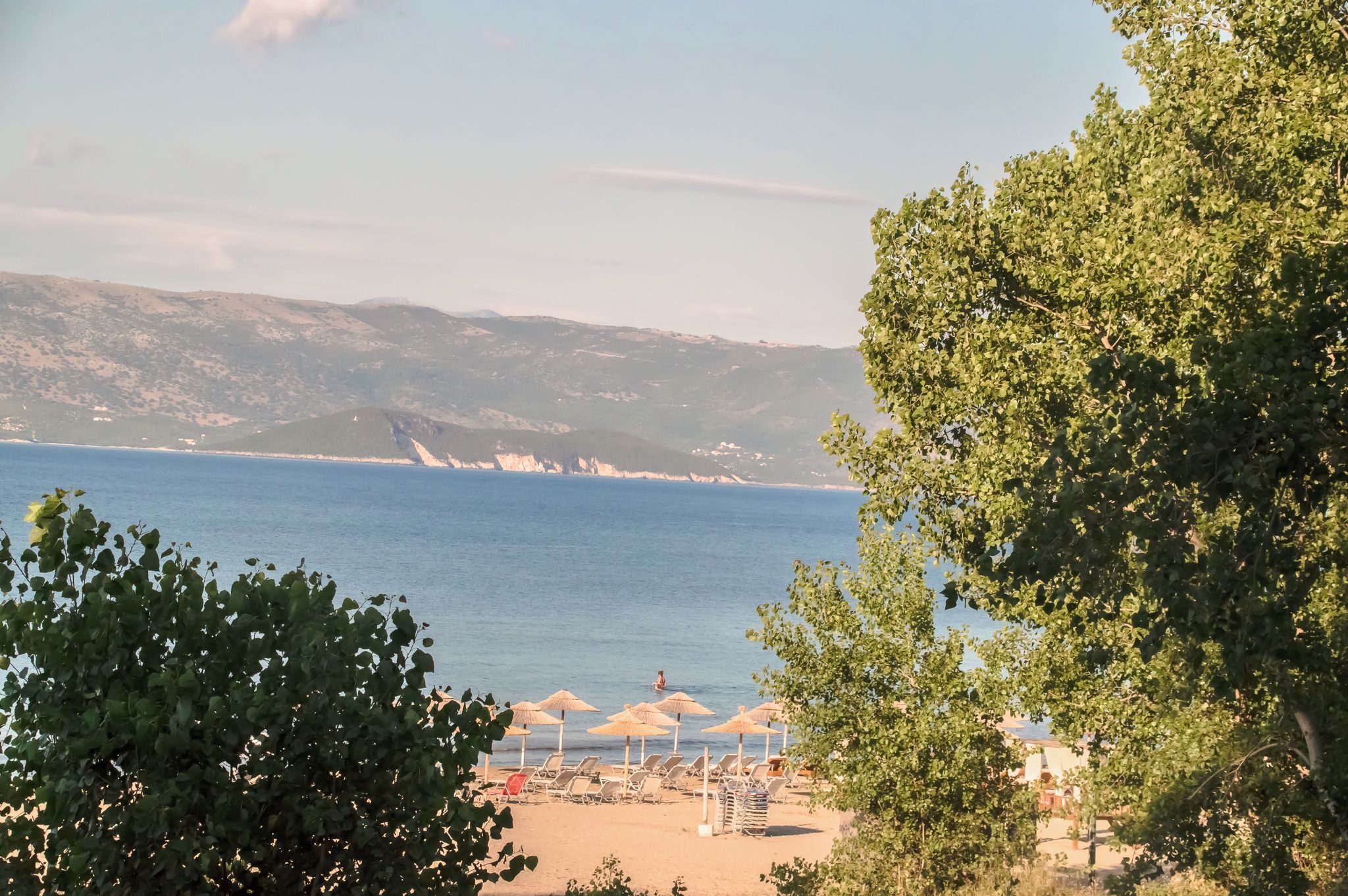 Una playa con sombrillas y sillas de descanso, vista despejada al mar y montañas a lo lejos.