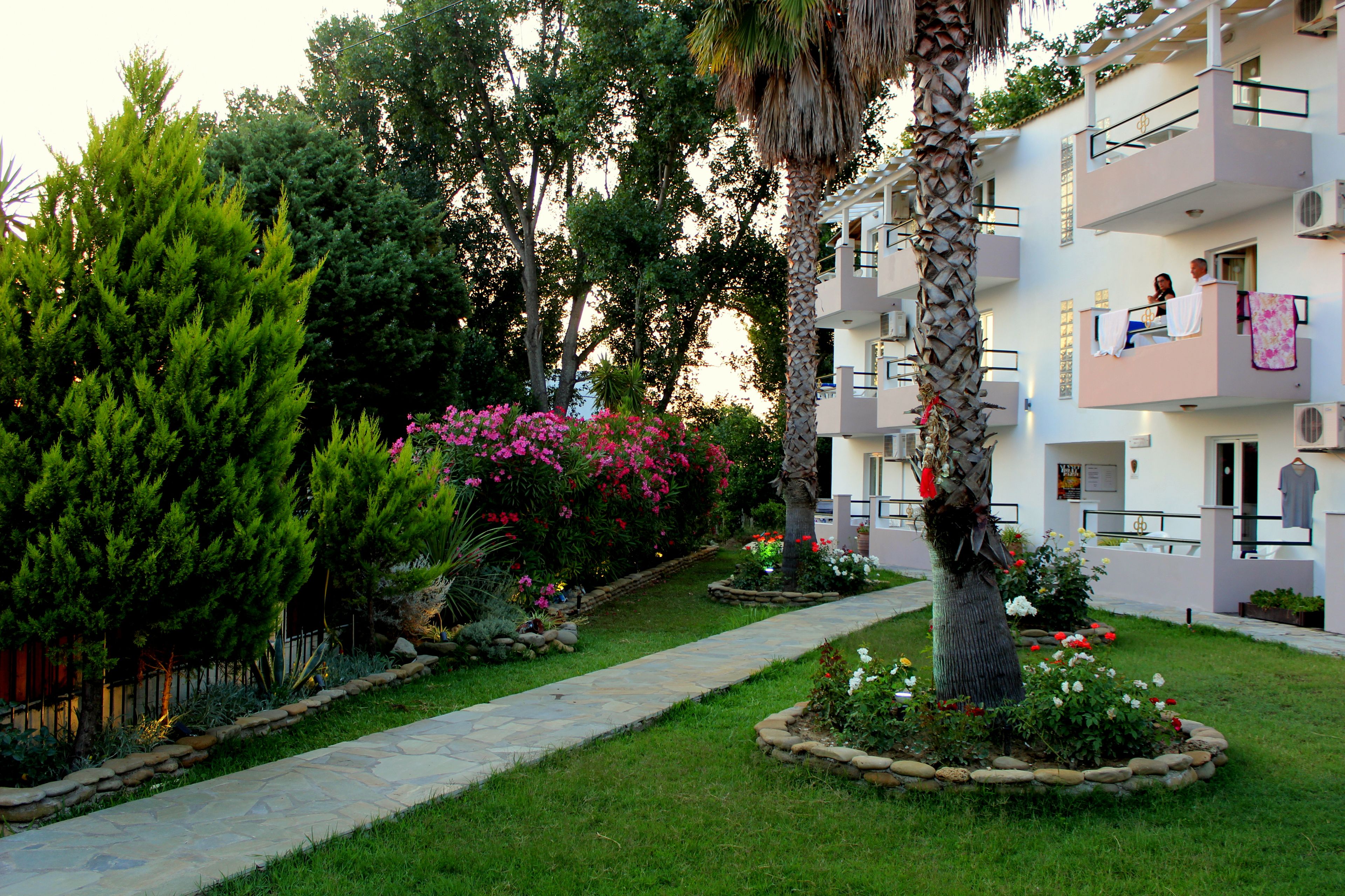 A garden pathway leading to a building with balconies, surrounded by greenery and flowering plants.