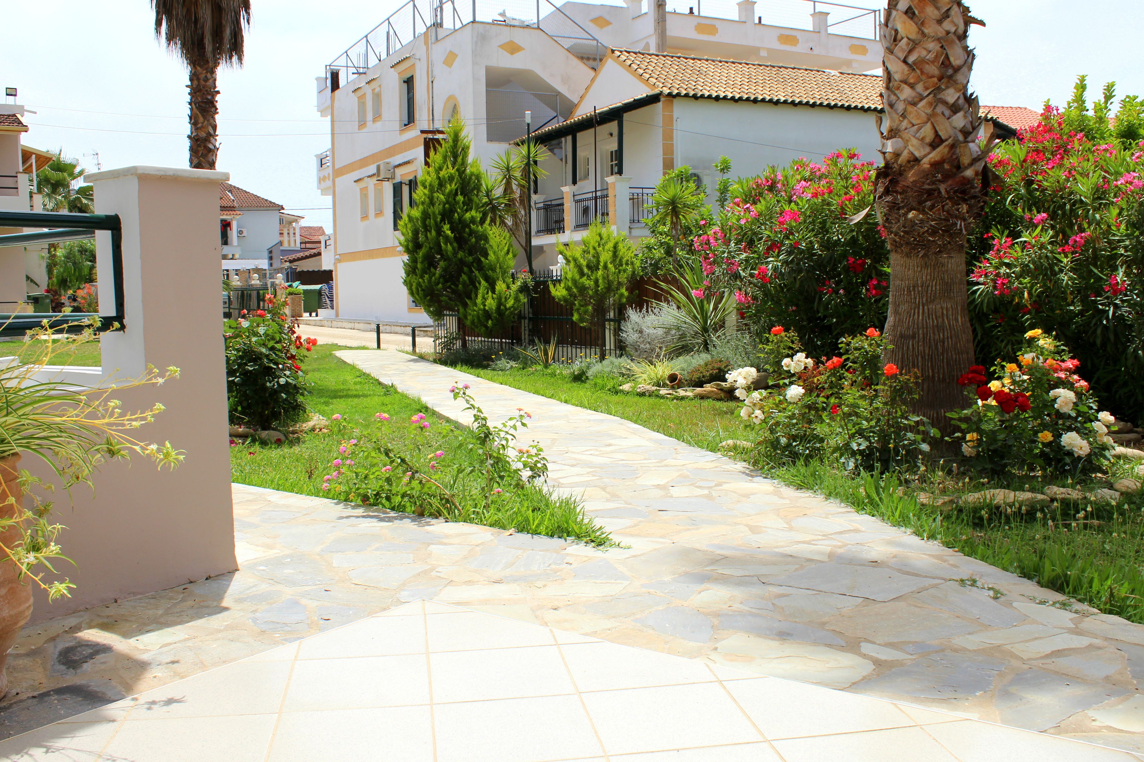 A pathway leading to a modern building with a balcony, surrounded by lush greenery and flowering plants.
