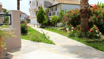 A pathway leading to a modern building with a balcony, surrounded by lush greenery and flowering plants.