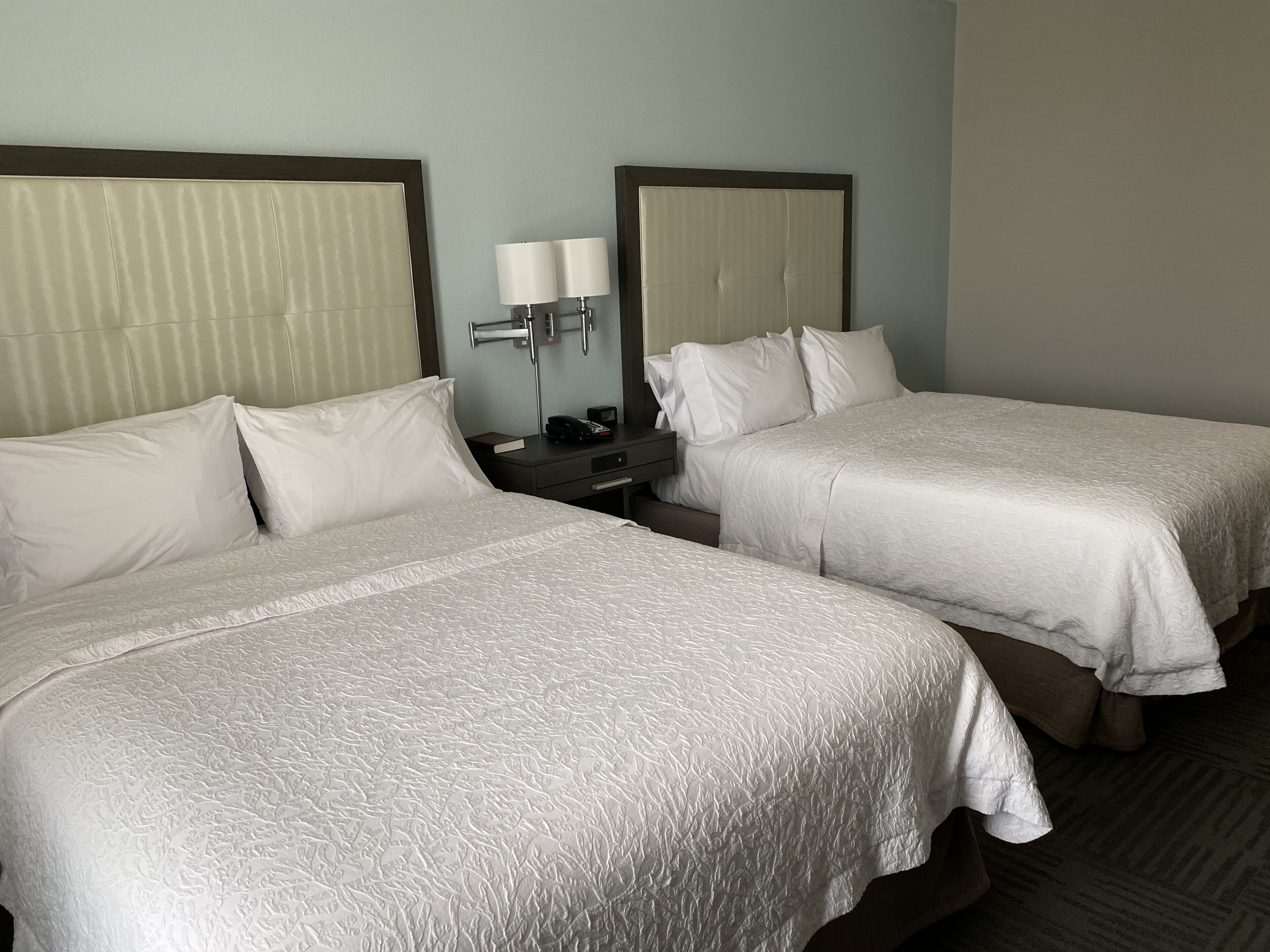A hotel room with two beds, each with a headboard and a bedside table with a lamp.