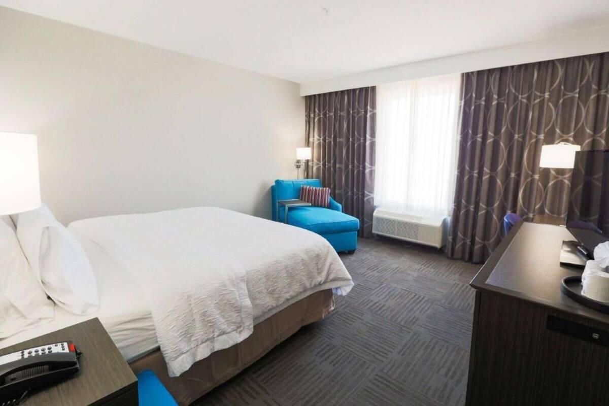 Ein Hotelzimmer mit Bett, Schreibtisch, Computer, Stuhl und einem Fenster mit Vorhängen.