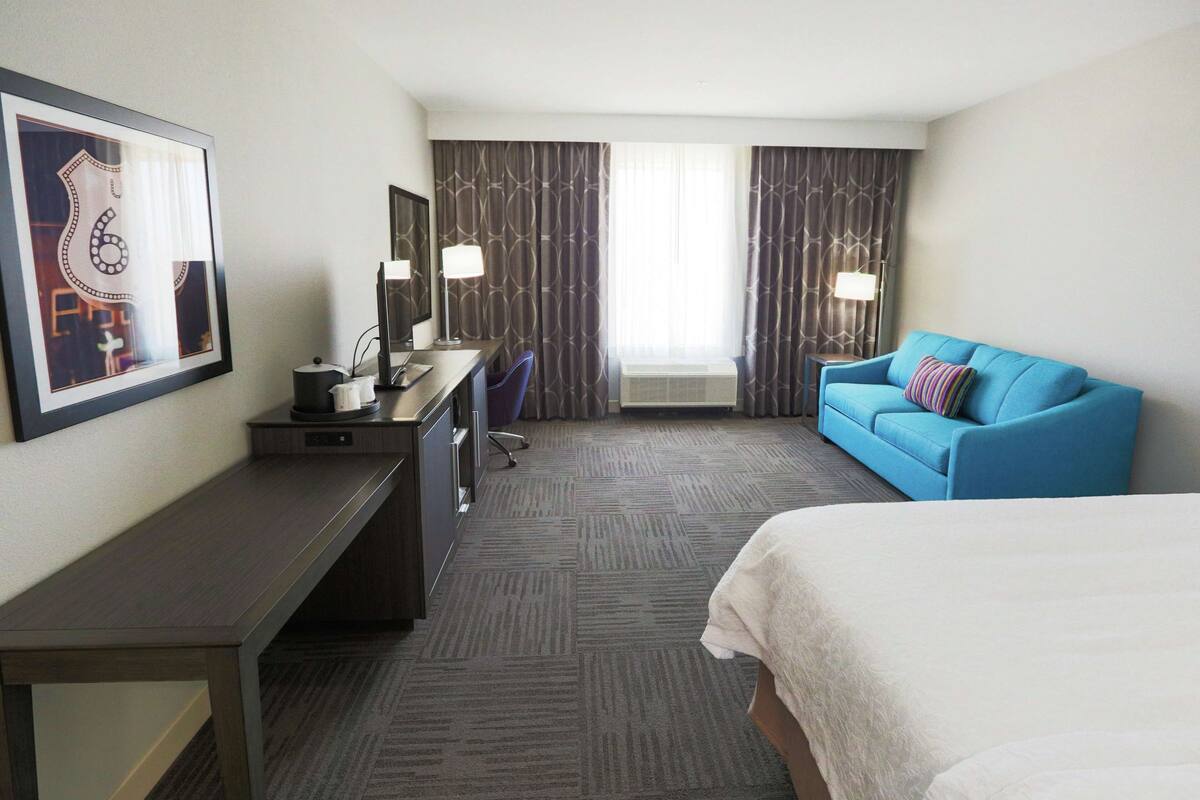 Ein Hotelzimmer mit einem Bett, einem Schreibtisch, einem Sessel, einem blauen Sofa und einem großen Fenster mit Vorhängen.