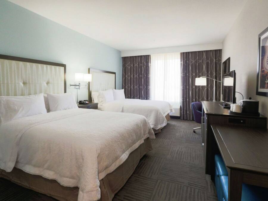 A hotel room with two beds, a desk, a chair, and a large window with curtains.