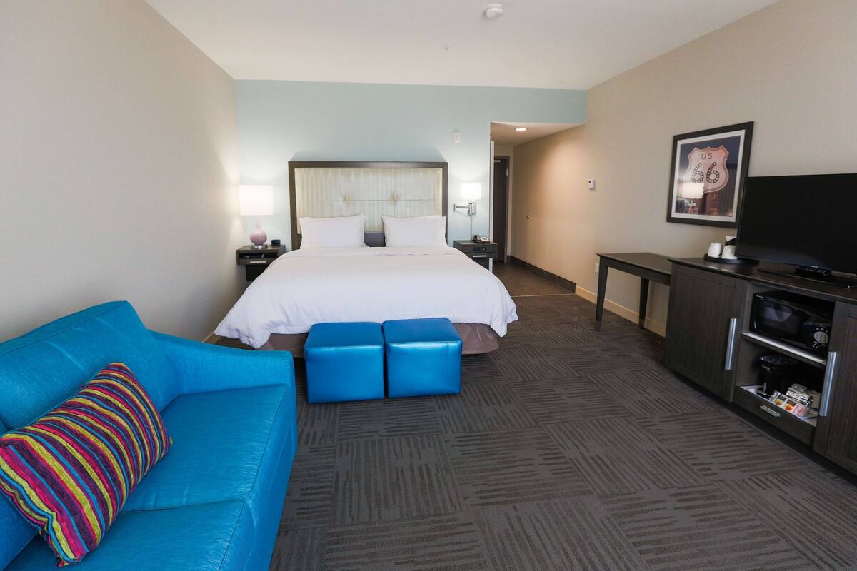 Ein Hotelzimmer mit einem großen Bett, einem blauen Sofa, einem farbenfrohen gestreiften Kissen, einem Fernseher und einer Minibar.