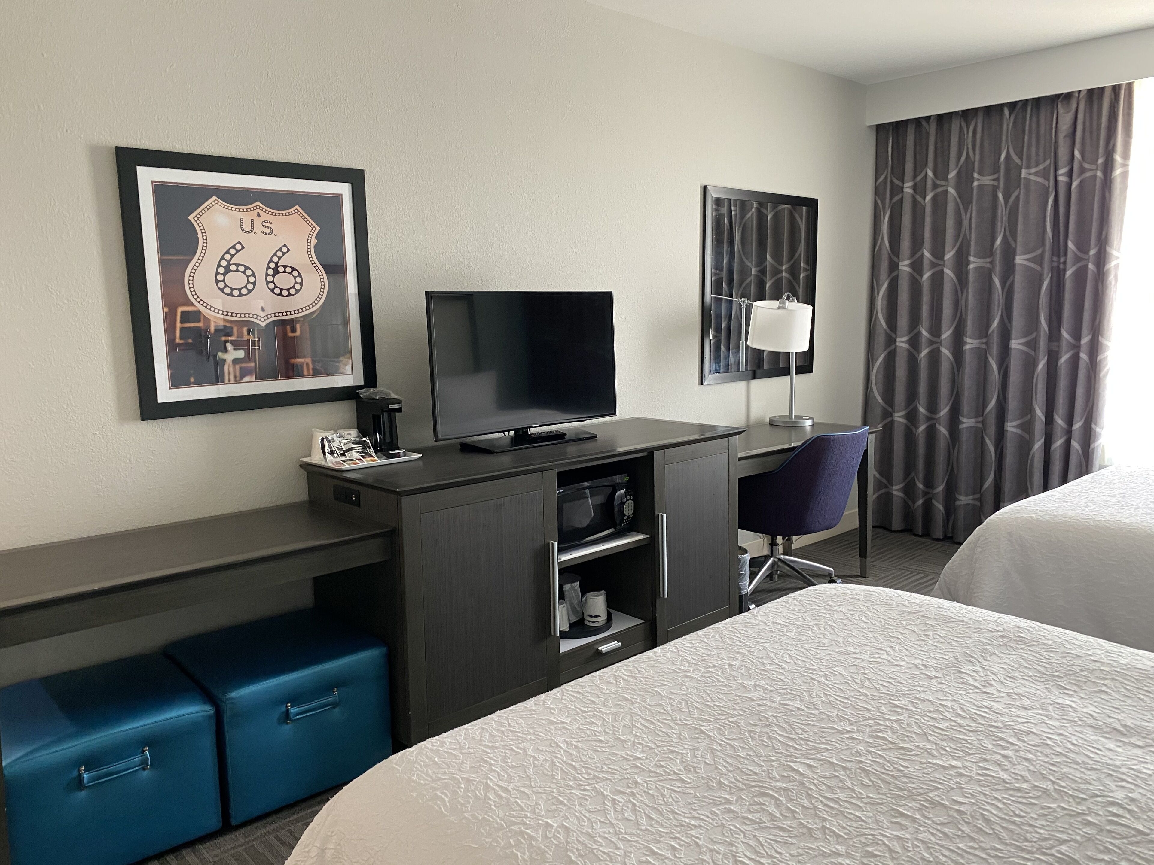 A hotel room with a bed, a desk with a computer, a TV, a coffee maker, and a framed picture of a road sign.