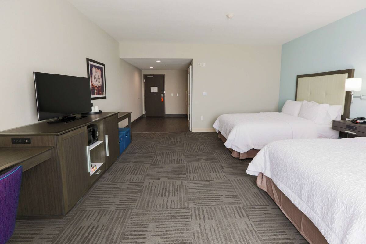 Ein Hotelzimmer mit Flachbildfernseher, Schreibtisch mit Computer, zwei Betten, Nachttisch und einer Wandlampe.
