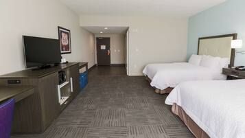A hotel room with a flat-screen TV, a desk with a computer, two beds, a nightstand, and a wall-mounted lamp.