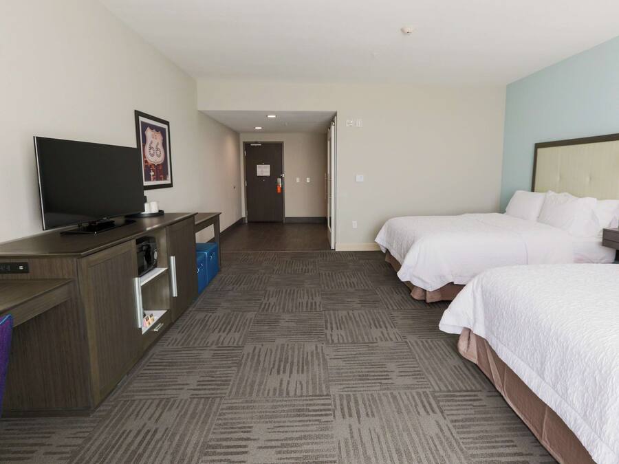 A hotel room with a flat-screen TV, a desk with a computer, two beds, a nightstand, and a wall-mounted lamp.