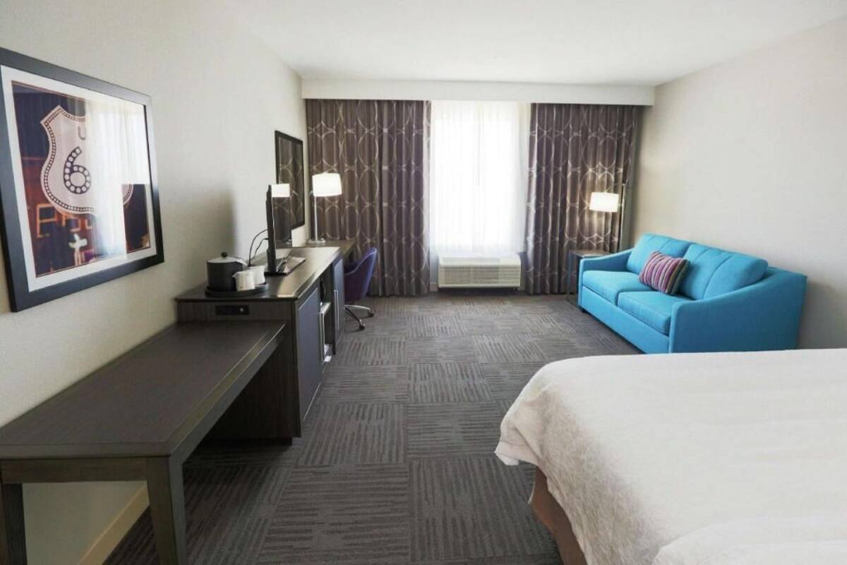 Ein Hotelzimmer mit einem Bett, einem Schreibtisch, einem Stuhl, einem blauen Sofa und einem großen Fenster mit Vorhängen.