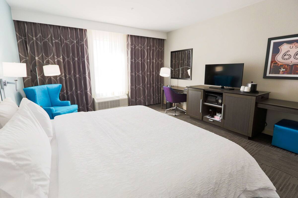 Ein Hotelzimmer mit einem großen Bett, einem Schreibtisch mit Fernseher, einem Sessel und einem großen Fenster mit Vorhängen.