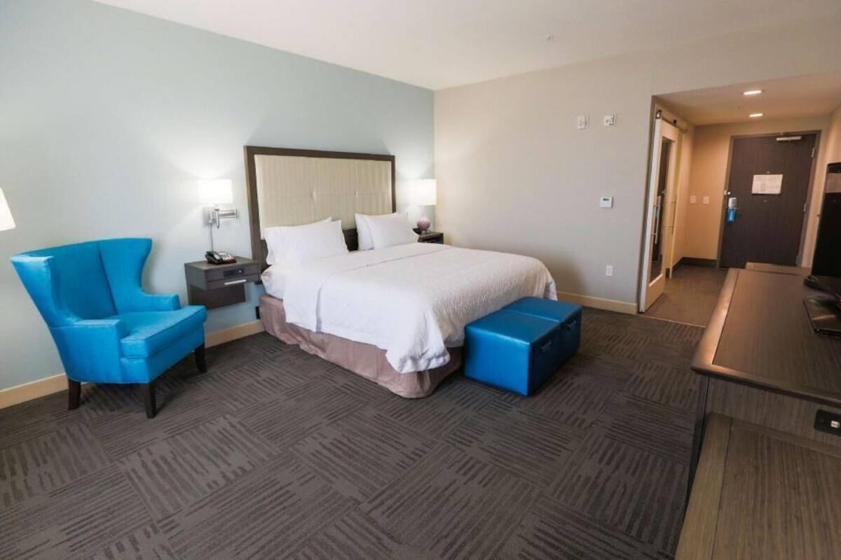 Ein Hotelzimmer mit einem Bett, einem blauen Sessel, einem Nachttisch, einem Schreibtisch und einer Tür.