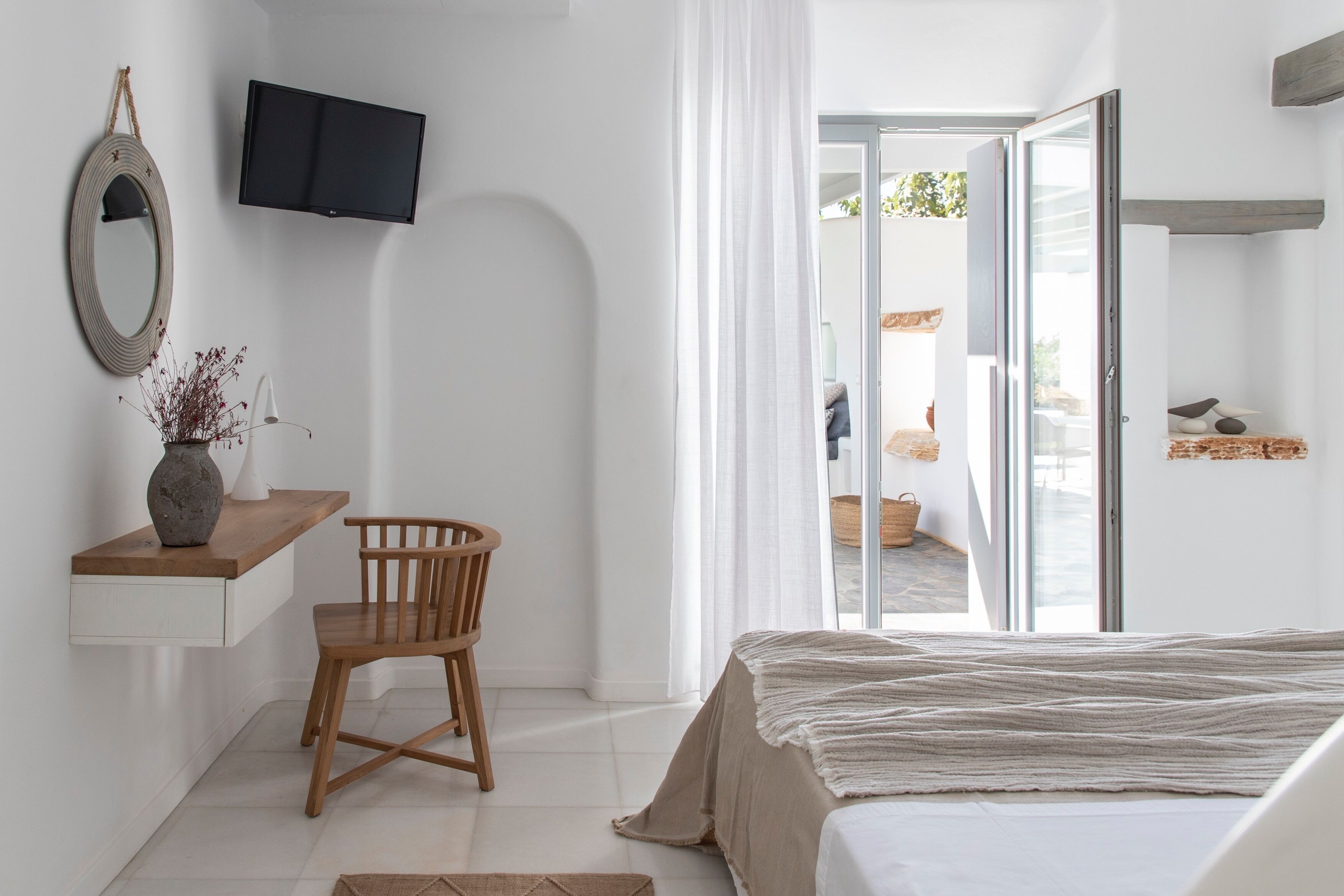 A bedroom with a bed, a wooden chair, a desk with a vase, a television, and a sliding glass door leading to an outdoor area.