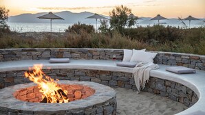 A stone fire pit surrounded by a curved seating area with cushions, overlooking a beach with umbrellas and a sunset view. - Aqua Breeze Villas Naxos (Naxos)