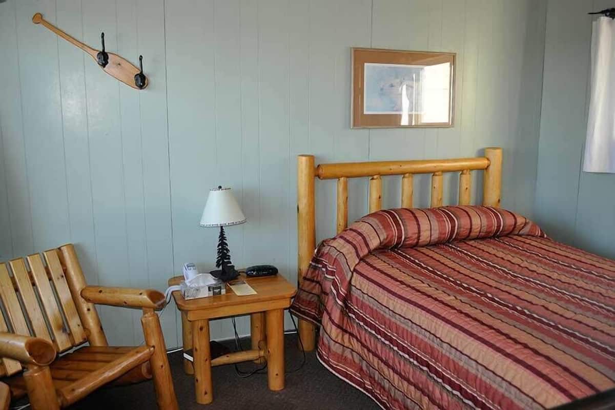 Een kamer met een houten bed, een houten stoel, een klein tafeltje met een lamp en een ingelijst schilderij aan de muur.