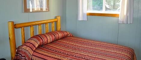 Una cama individual con cabecera de madera y colcha a rayas en una habitación con paredes azul claro y una ventana con cortinas blancas.