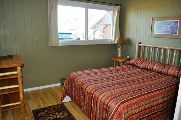 A bedroom with a wooden bed, a nightstand, a lamp, and a window with a view of a building outside.