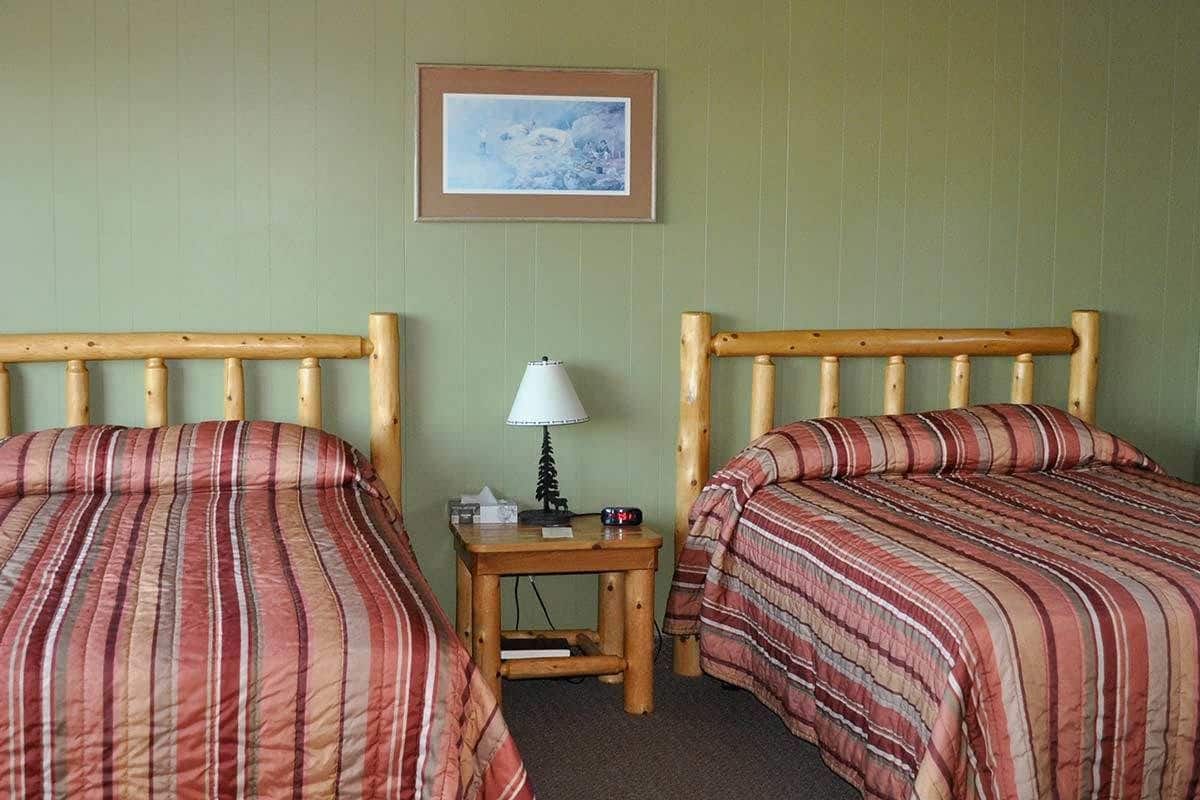 Een kamer met twee houten bedden, een kleine tafel met een lamp, en een lijst met een afbeelding aan de muur.