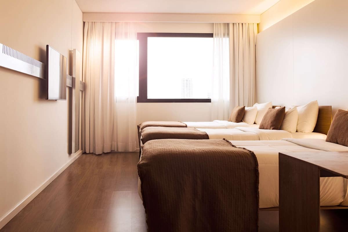 Une chambre d’hôtel avec deux lits, une grande fenêtre avec des rideaux, un téléviseur à écran plat fixé au mur et un plancher en bois.