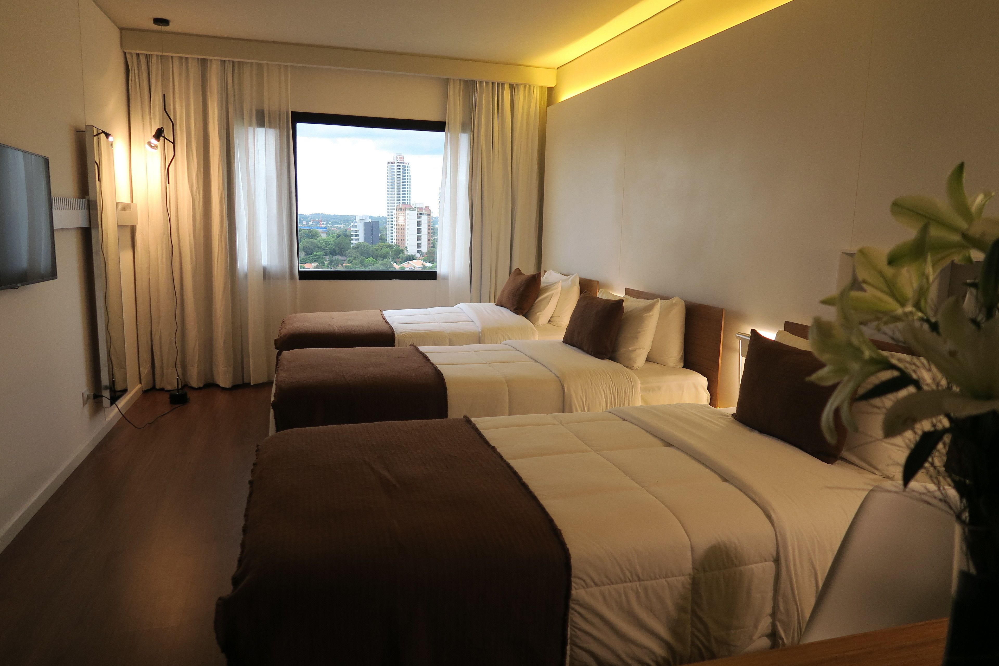 A hotel room with three beds, a large window with a city view, and a flat-screen TV mounted on the wall.