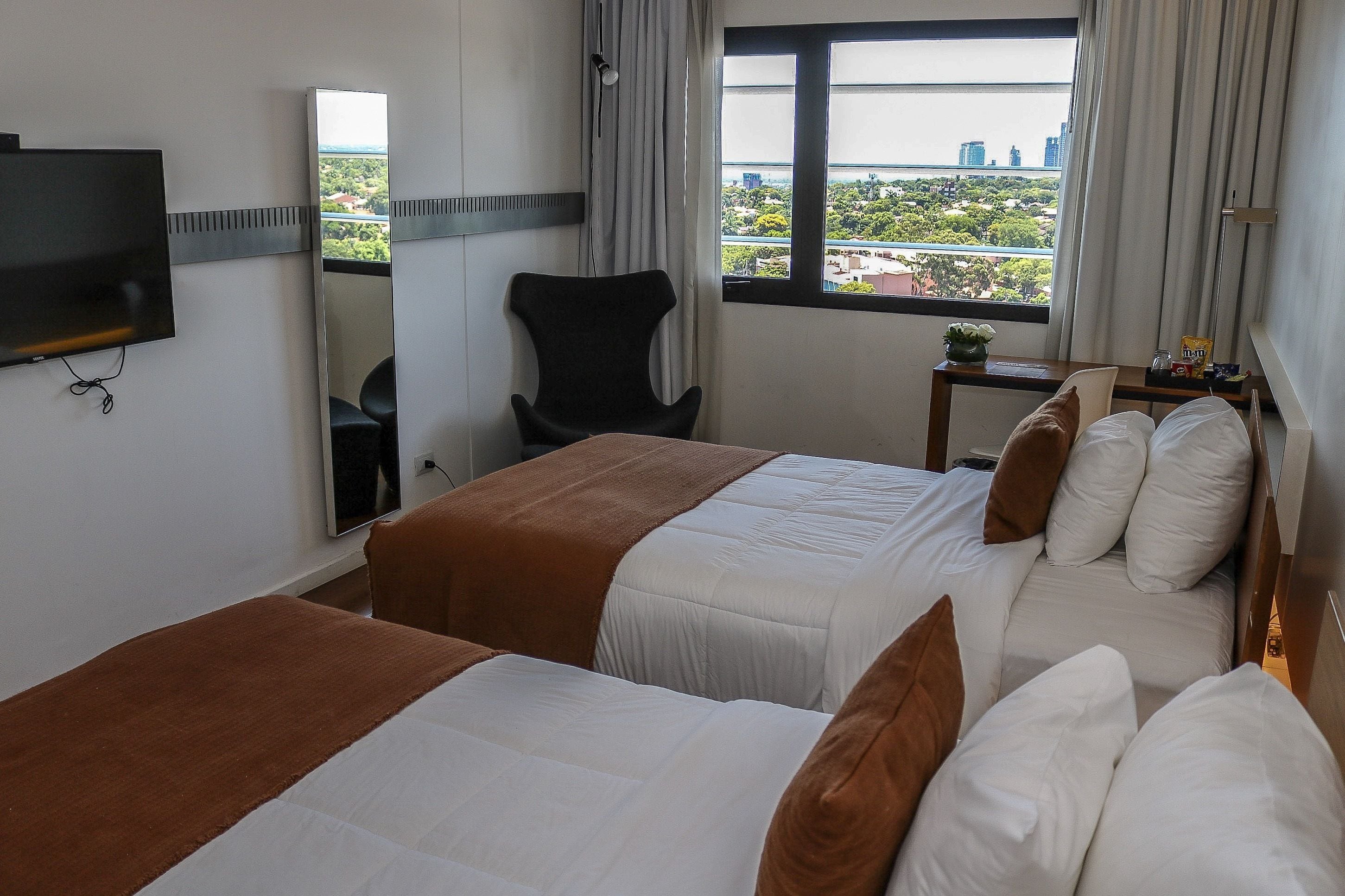 A hotel room with two beds, a TV, a chair, a small table, and a window with a city view.