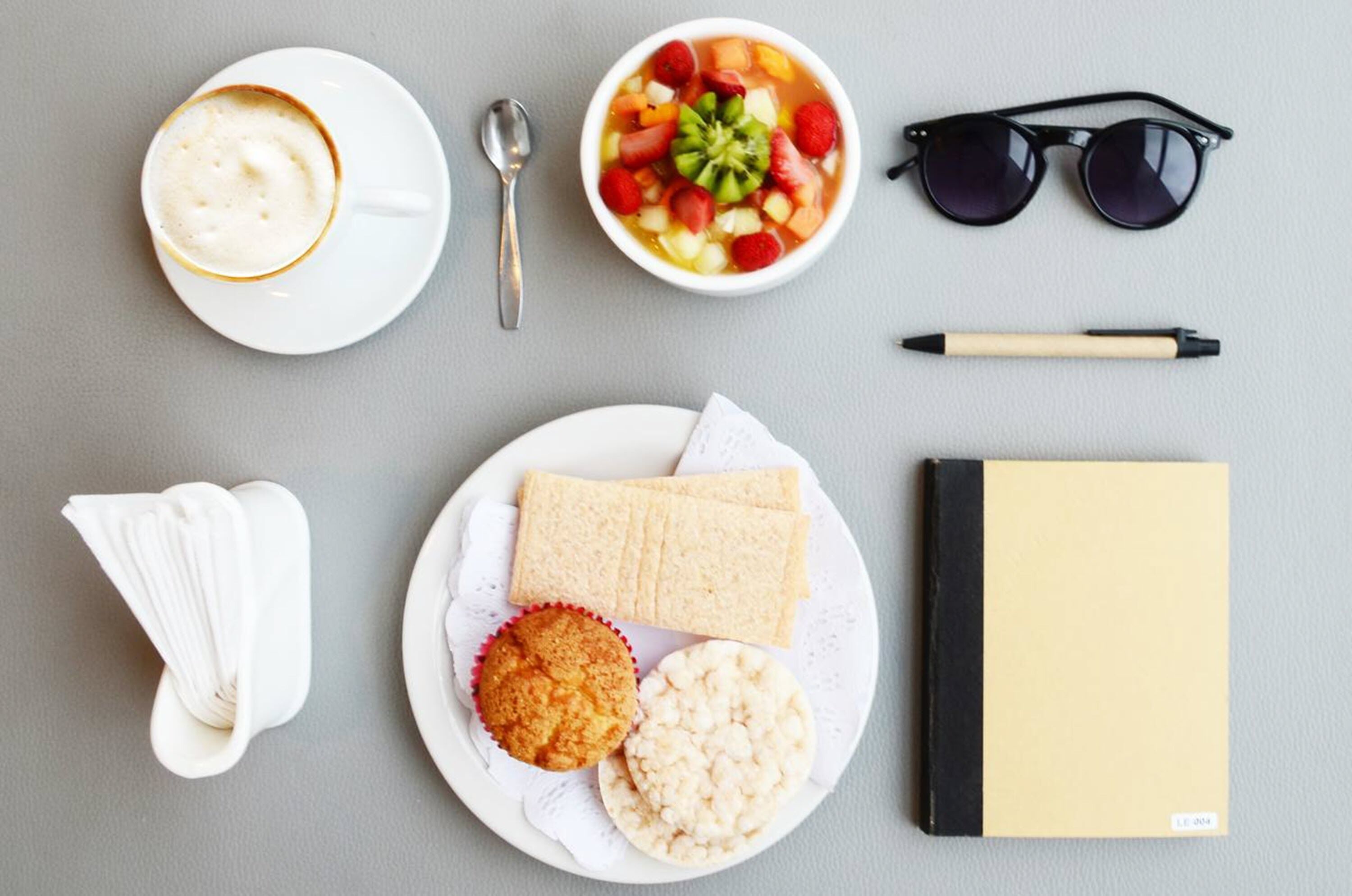 Ein Frühstücksangebot mit einer Tasse Kaffee, einer Obstschale, einem Muffin, Keksen, einem Sandwich, einem Notizbuch, einem Stift, einer Sonnenbrille und einem Löffel.