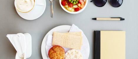Ein Frühstücksangebot mit einer Tasse Kaffee, einer Obstschale, einem Muffin, Keksen, einem Sandwich, einem Notizbuch, einem Stift, einer Sonnenbrille und einem Löffel.