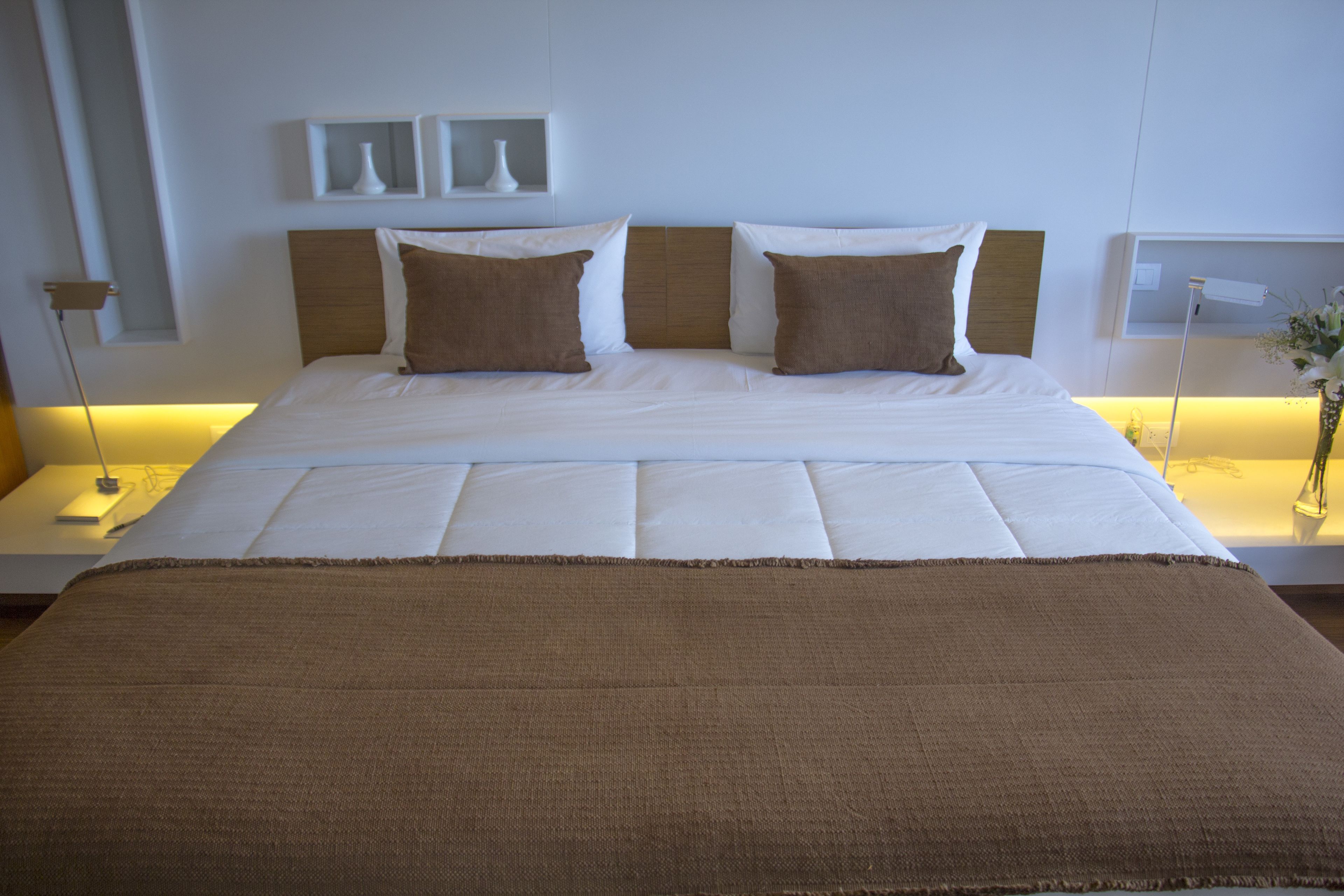A neatly made bed with a brown blanket and two pillows, a bedside table with a lamp, and a wall-mounted shelf with decorative items.