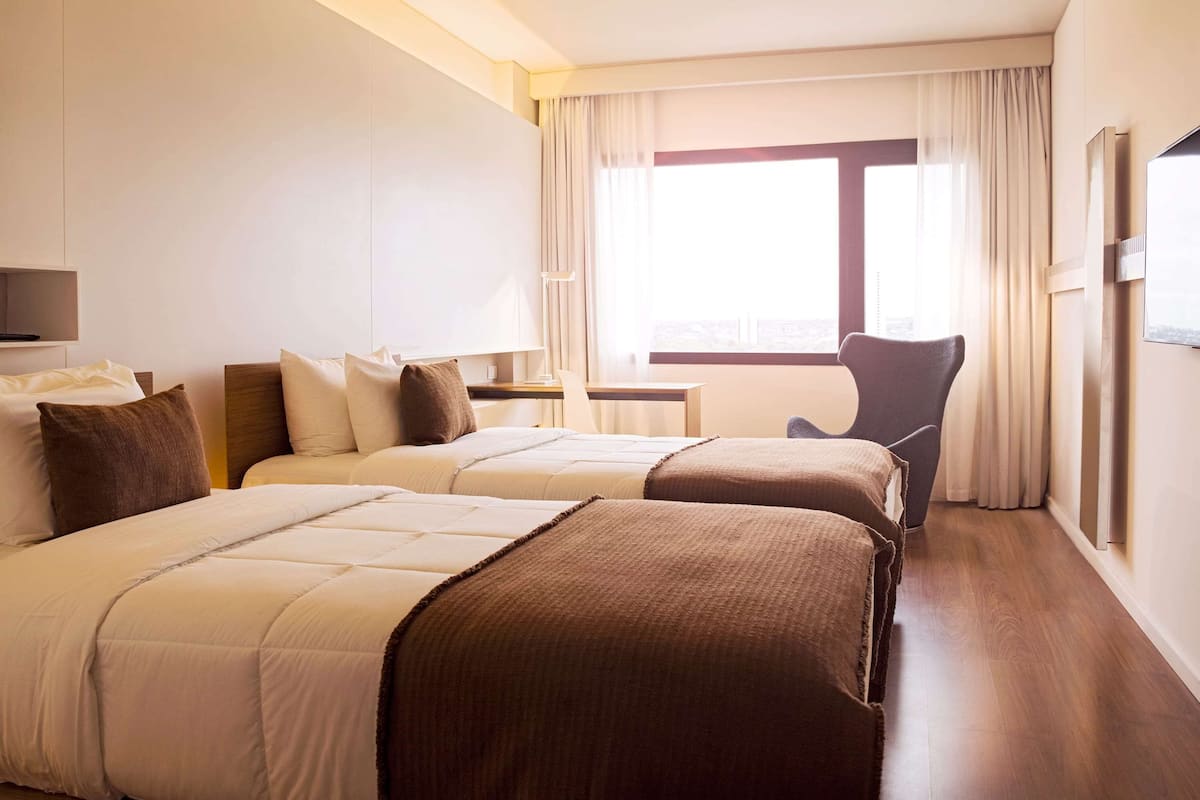 Une chambre d’hôtel avec deux lits, un bureau, une chaise et une grande fenêtre avec des rideaux.