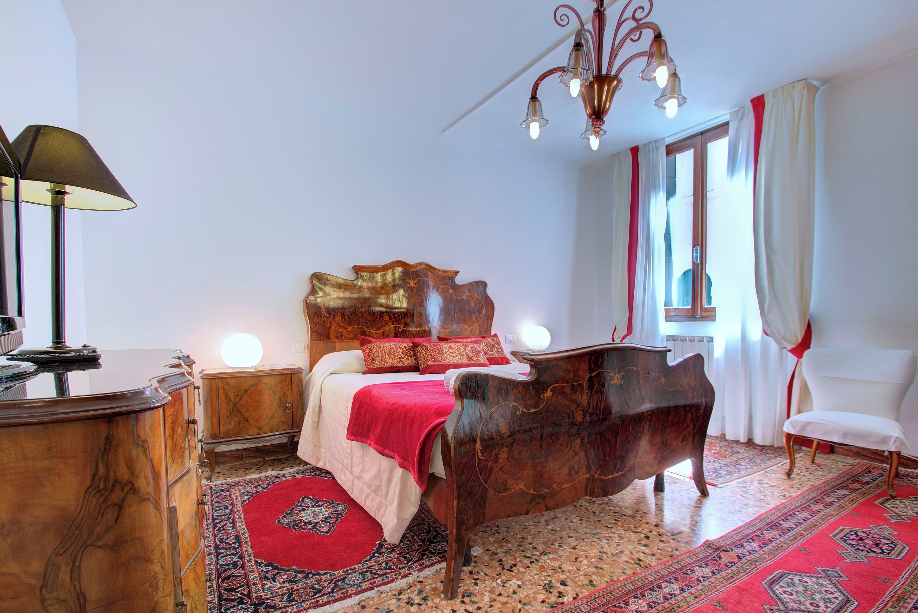A bedroom with a wooden bed, a nightstand, a lamp, a chandelier, and a red and beige rug.