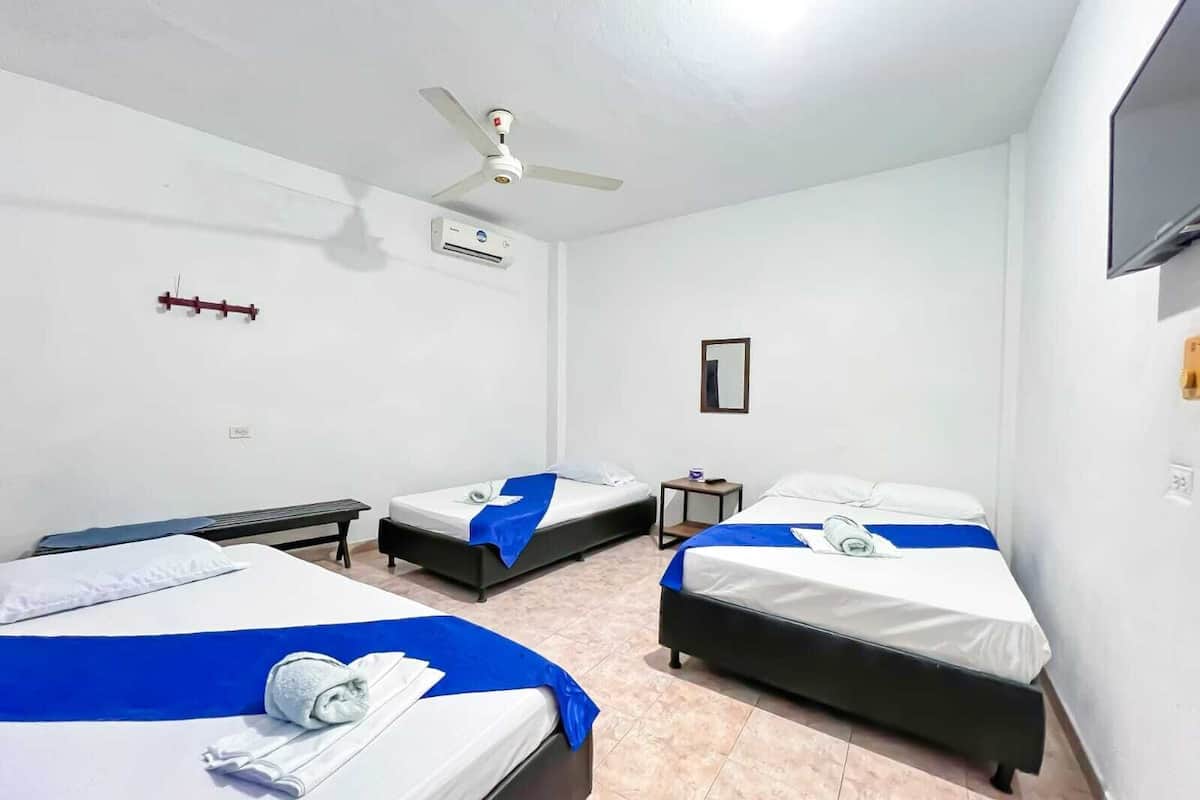 A hotel room with three beds, a ceiling fan, an air conditioning unit, a small table, and a television mounted on the wall.