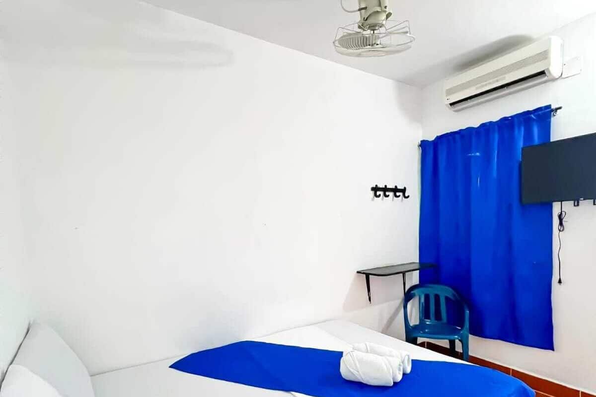 A single-bed room with white bedding, a blue accent on the bed, a ceiling fan, an air conditioning unit, a blue curtain, a small table with a chair, and a TV mounted on the wall.