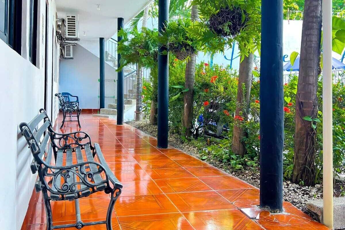 A covered corridor with a bench, air conditioning units, and a garden with palm trees and plants.