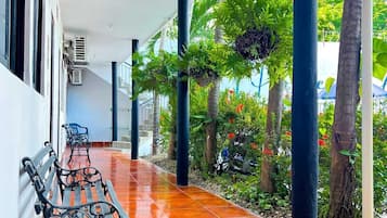 Un corredor cubierto con un banco, unidades de aire acondicionado y un jardín con palmeras y plantas.