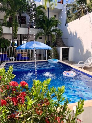 A swimming pool with a water fountain, surrounded by palm trees and a blue umbrella. - Hotel Zulan (Tolú)