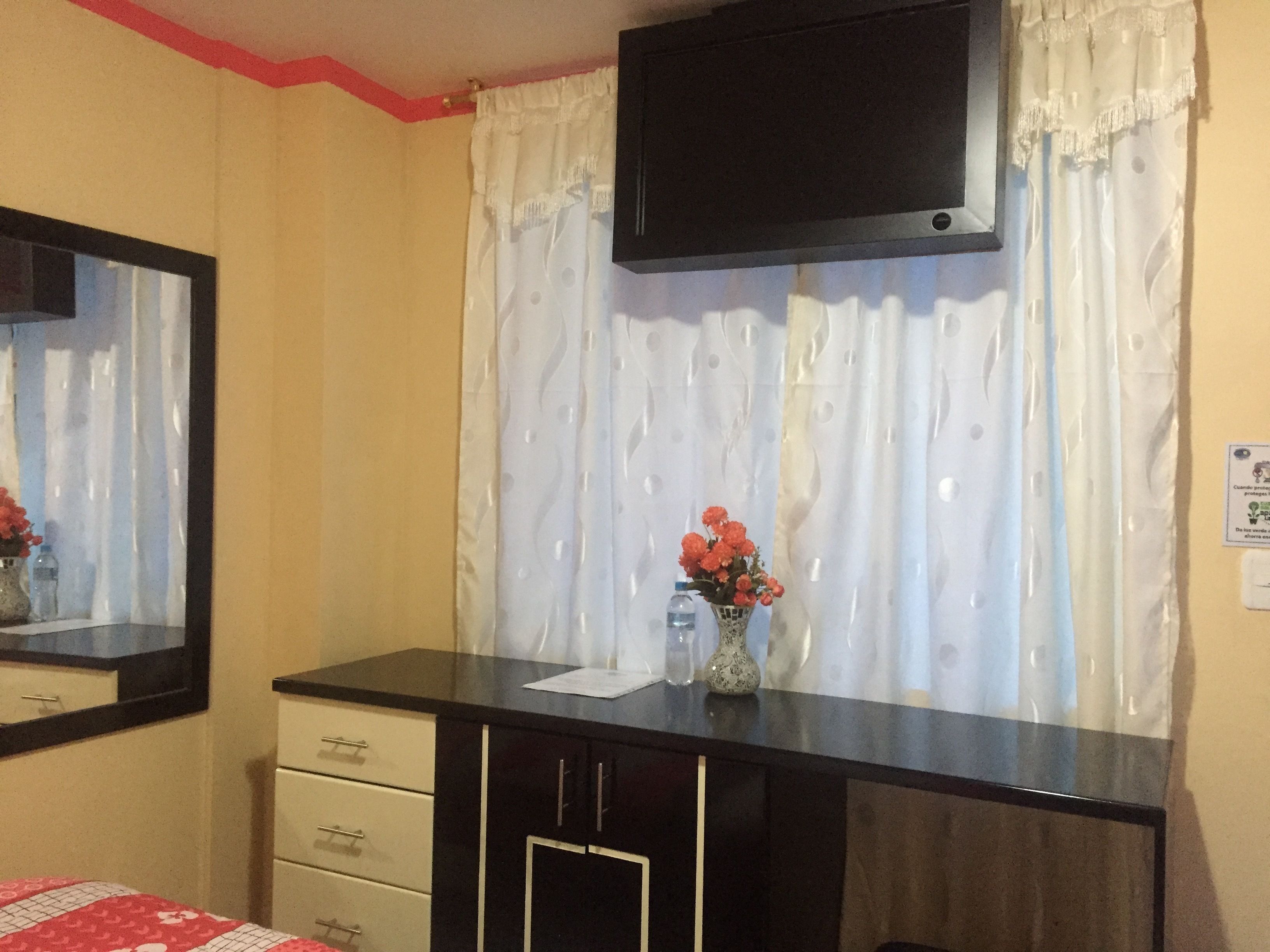 A bedroom with a bed, a dresser, a TV mounted on the wall, and a window with curtains.