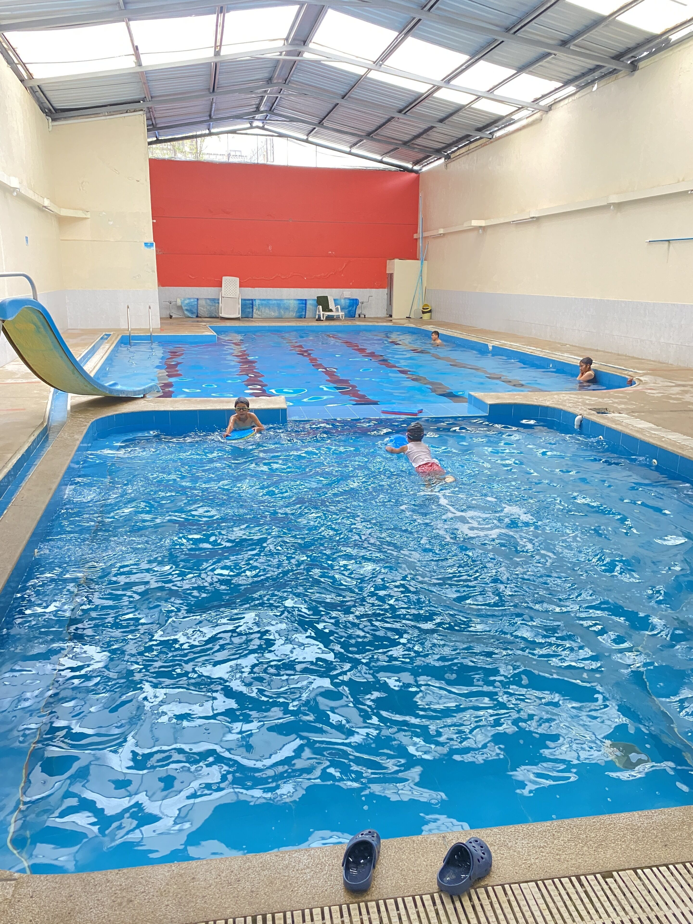An indoor swimming pool with multiple lanes, a diving board, and a red wall in the background.
