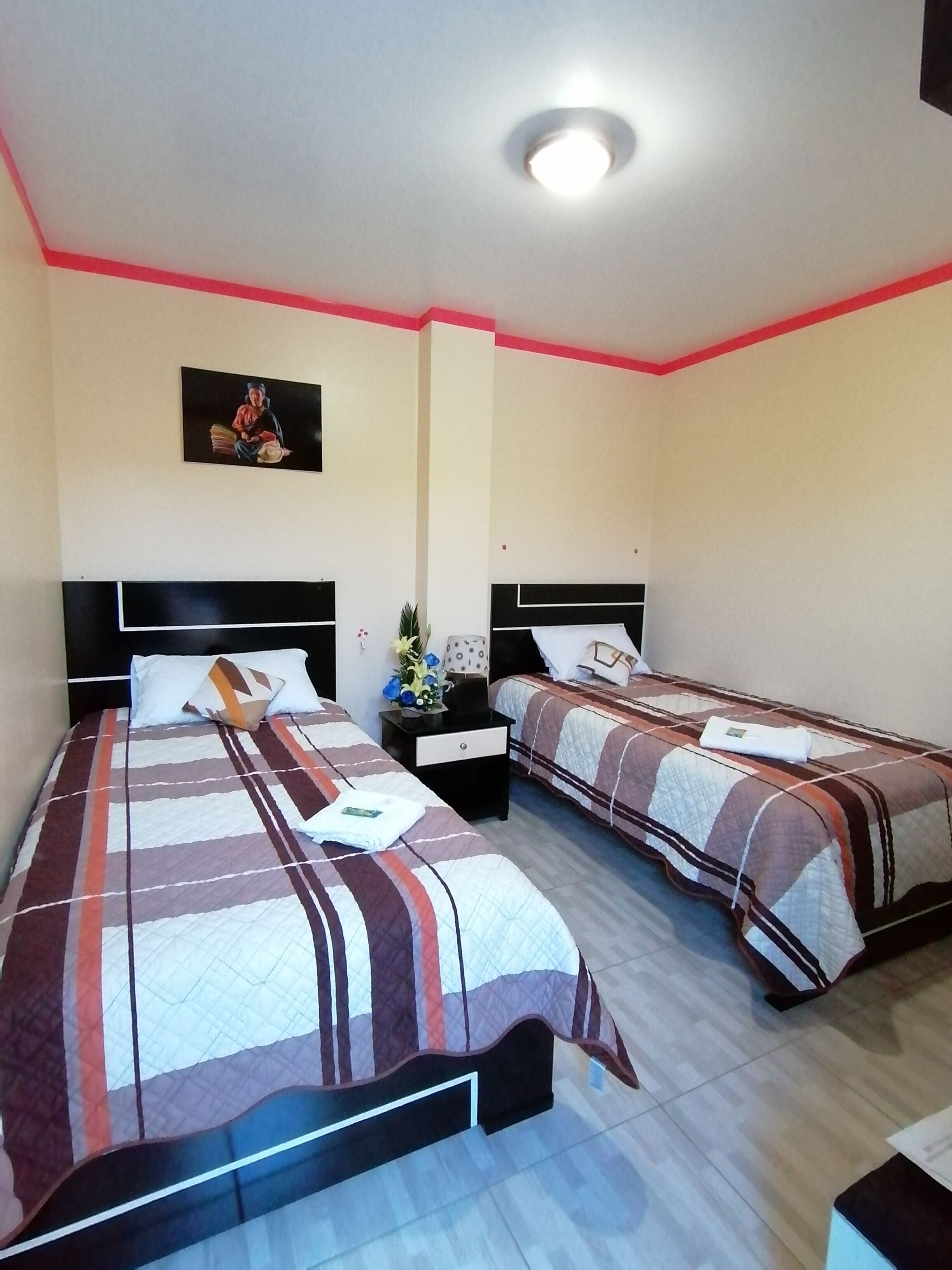 Habitación de hotel con dos camas, cada una con una colcha a rayas, una mesita de noche con lámpara y un cuadro en la pared.