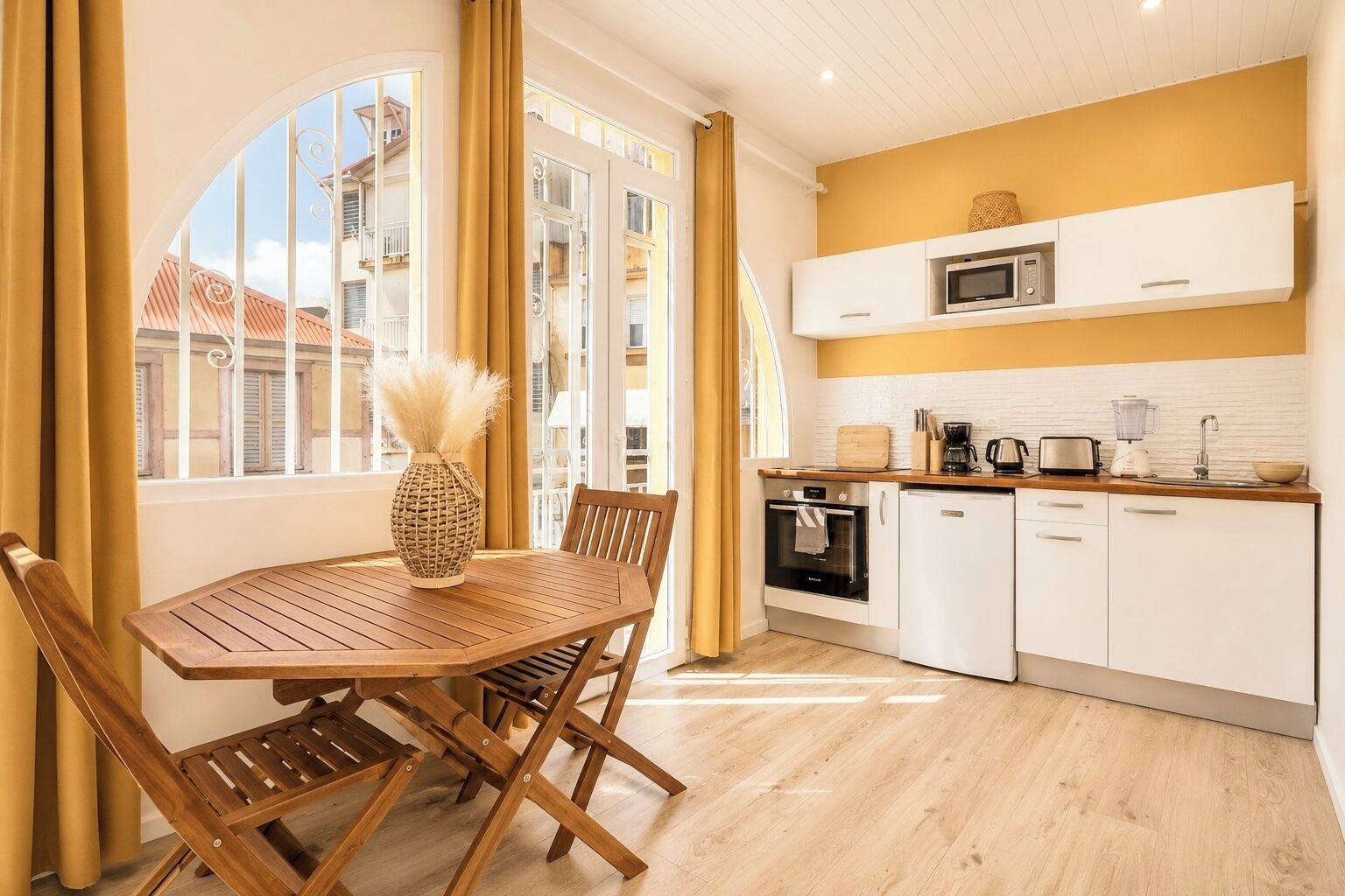 A compact kitchen with a dining area, featuring a wooden table and chairs, a built-in oven, microwave, and white cabinetry.