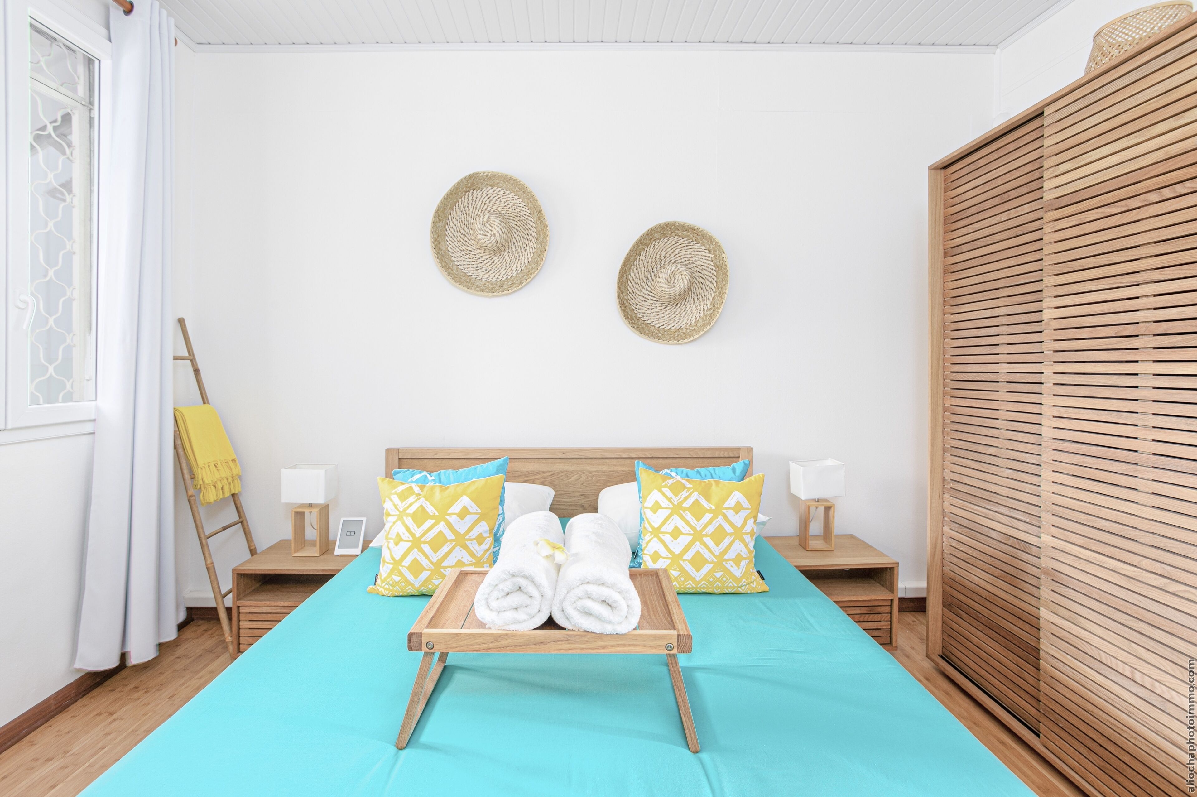 A neatly arranged hotel room with a bed, two wall-mounted woven baskets, a wooden ladder, and a wooden wardrobe.