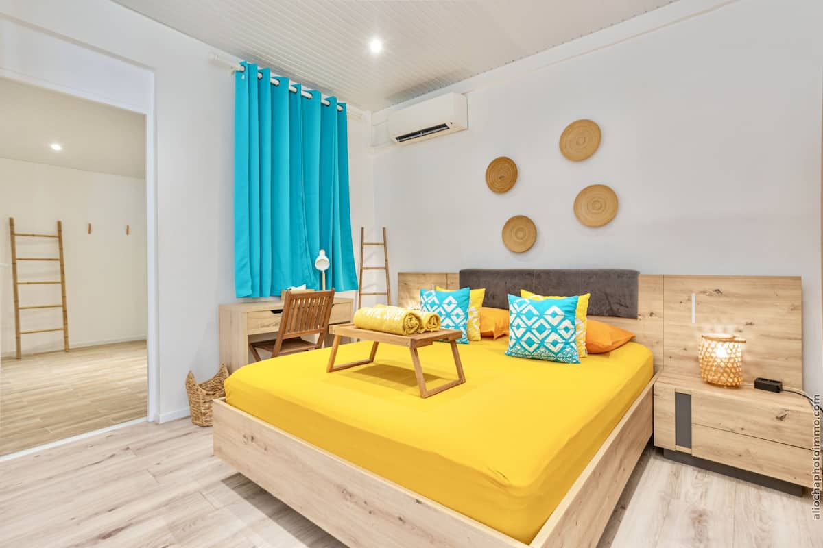 A modern bedroom with a wooden bed, bright yellow bedding, a wooden nightstand, a desk with a chair, and a window with blue curtains.