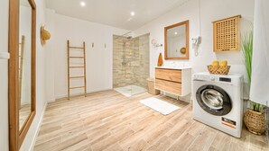 A modern bathroom with a wooden floor, a glass shower enclosure, a washing machine, and a wooden vanity with a mirror. - Foyal Suites (Fort-de-France)