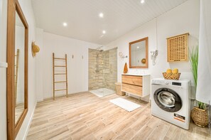 A modern bathroom with a wooden floor, a glass shower enclosure, a washing machine, and a wooden vanity with a mirror. - Foyal Suites (Fort-de-France)