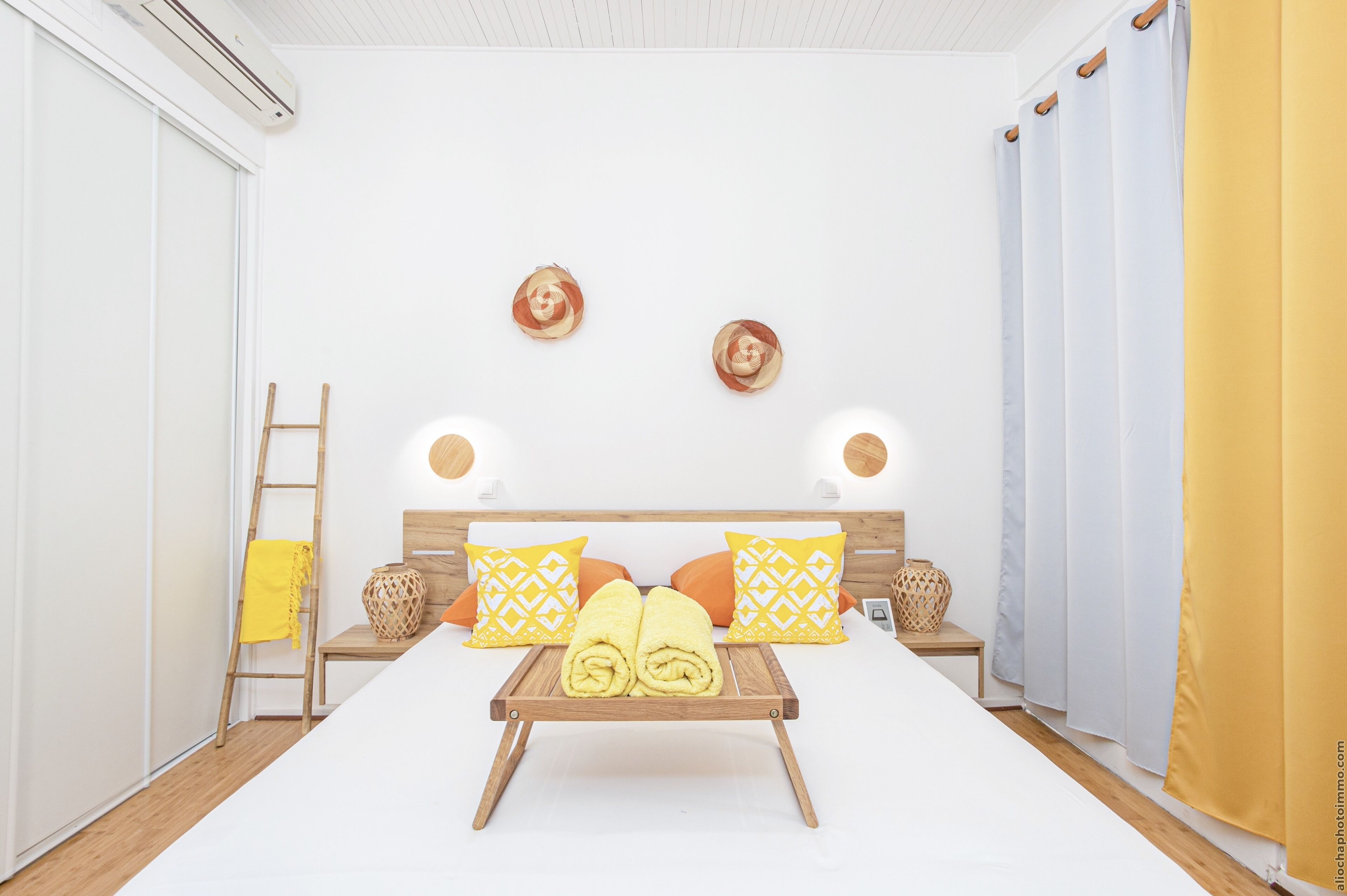 Habitación de hotel con cama, mesita de noche, lámpara, bandeja con toallas y ventana con cortinas.