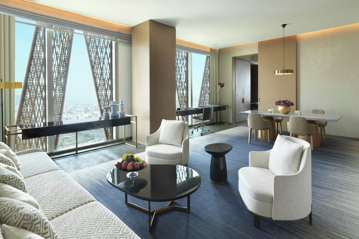 Une chambre d’hôtel moderne dotée d’un grand canapé, de deux fauteuils, d’un coin repas avec une table et des chaises, et de grandes fenêtres offrant une vue sur la ville.