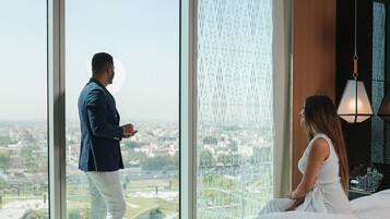 Ein Hotelzimmer mit großem Fenster und Blick auf die Stadt, einem Bett mit weißer Bettwäsche und einer Hängelampe.