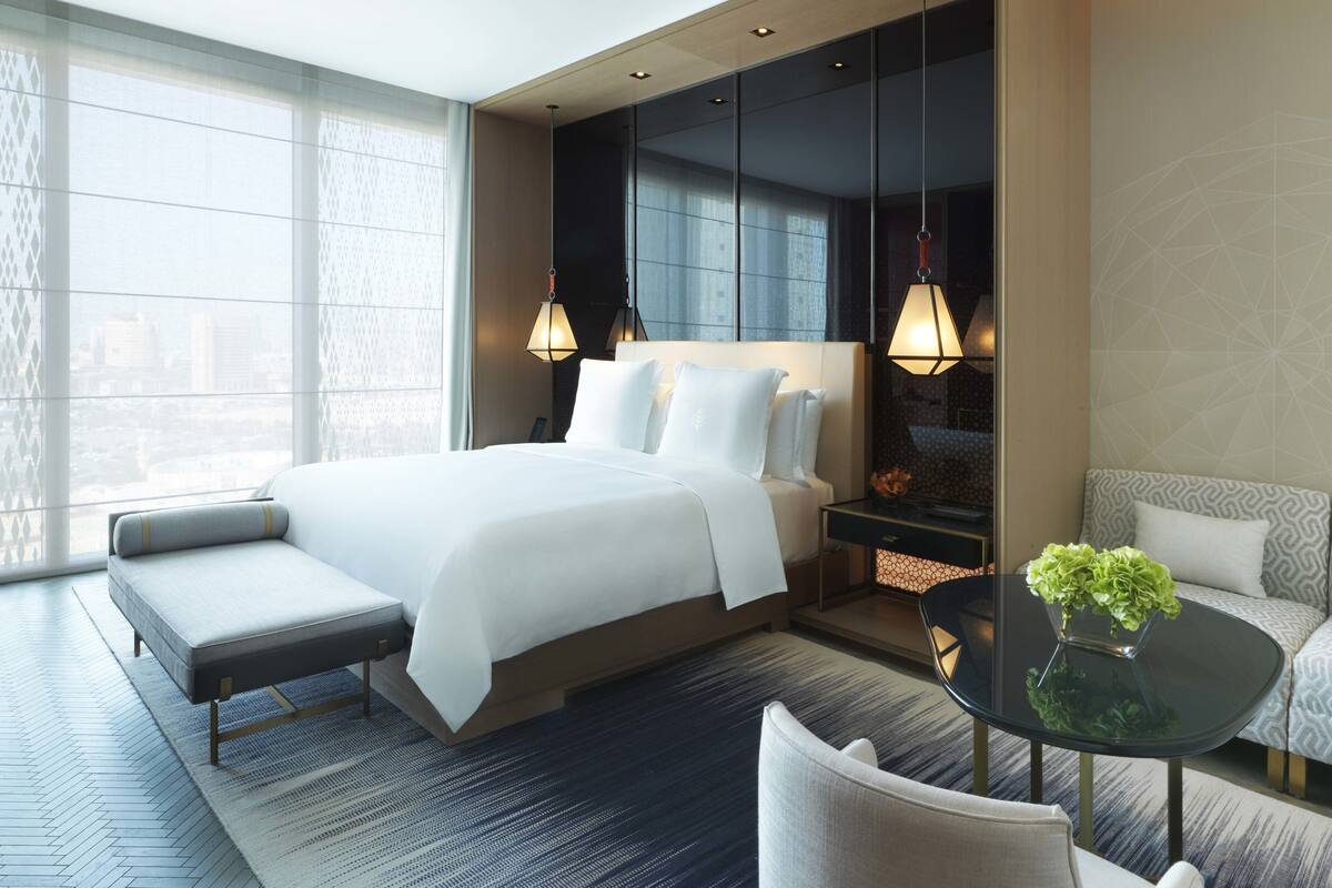 Une chambre d’hôtel moderne avec un grand lit, un canapé, une petite table avec une plante et une vue sur la ville grâce à de grandes fenêtres.